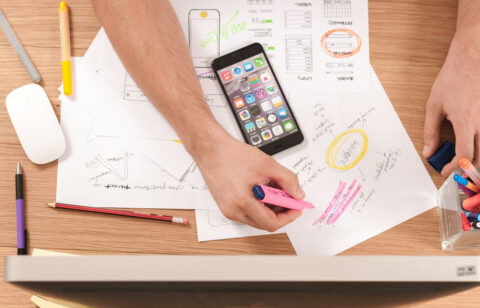 Person highlighting notes on wireframe sketches at a cluttered desk with a smartphone and colorful pens.