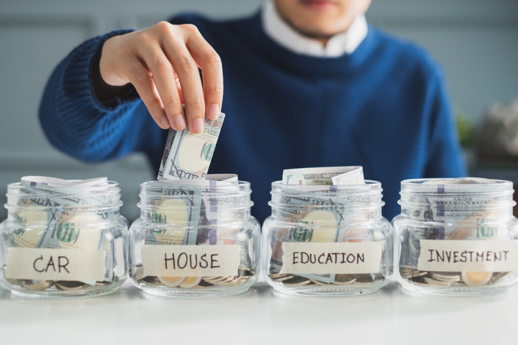 Person placing cash into a labeled savings jar for a house, alongside jars for a car, education, and investment.