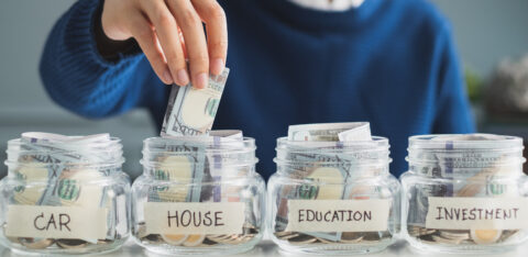 Person placing cash into a labeled savings jar for a house, alongside jars for a car, education, and investment.
