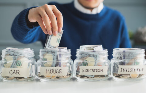 Person placing cash into a labeled savings jar for a house, alongside jars for a car, education, and investment.