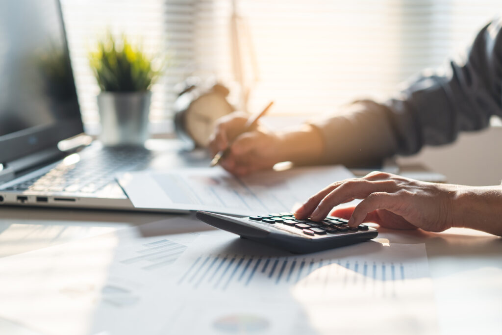 Person using a calculator and reviewing financial documents at a desk, with a laptop and charts in a sunlit office.