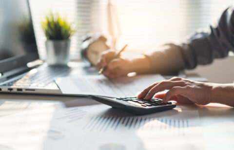 Person using a calculator and reviewing financial documents at a desk, with a laptop and charts in a sunlit office.