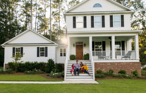 Family sitting on the front steps of a white suburban home with black shutters, enjoying time together outdoors.