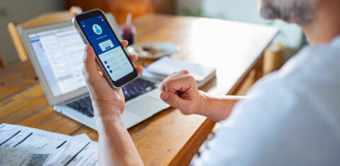Man checking his bank account on a smartphone while reviewing bills and using a laptop at home.