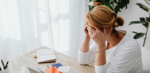 Stressed woman sitting at a desk with her head in her hands, surrounded by receipts, bills, and a calculator.