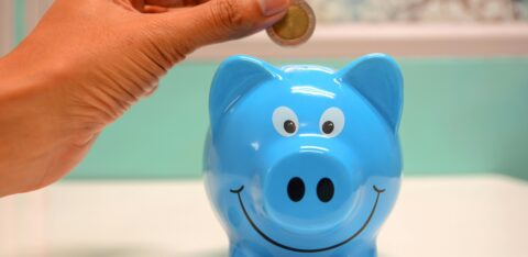 Smiling piggy bank with coins nearby and a hand about to add another coin.