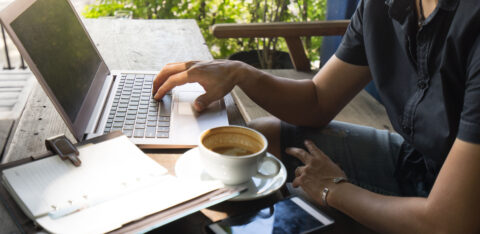 Person working on a laptop at an outdoor café table with a cup of coffee, a smartphone, and a notebook nearby.
