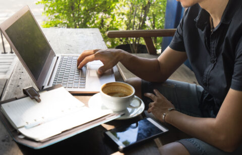 Person working on a laptop at an outdoor café table with a cup of coffee, a smartphone, and a notebook nearby.