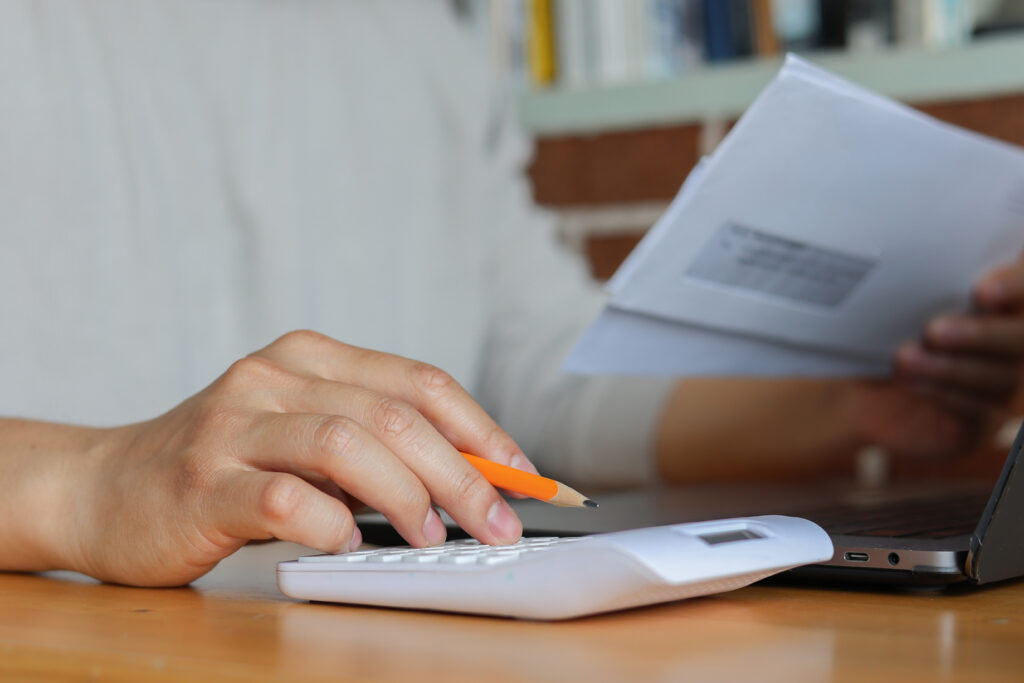 Close-up of hands using a white calculator and holding an envelope, with a pencil and laptop on a wooden desk.