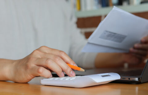 Close-up of hands using a white calculator and holding an envelope, with a pencil and laptop on a wooden desk.