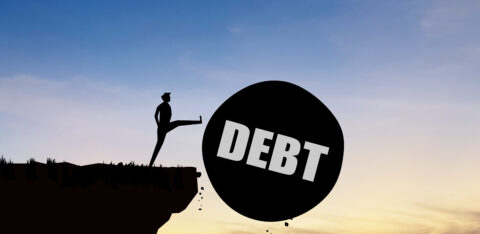 Silhouette of a person kicking a large ball labeled "DEBT" off a cliff at sunset.