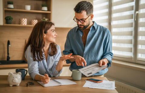 Couple reviewing household bills and budgeting together at a kitchen table with paperwork, coffee mugs, and a piggy bank nearby.