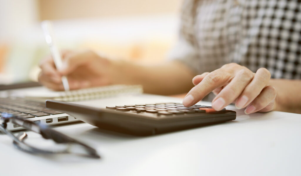 Close-up of a person using a calculator while taking notes next to a laptop on a desk.