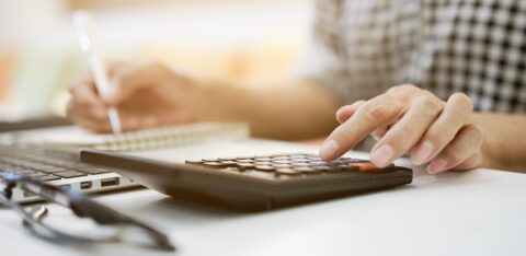 Close-up of a person using a calculator while taking notes next to a laptop on a desk.