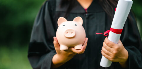 Graduate in a cap and gown holding a piggy bank and a diploma tied with a red ribbon