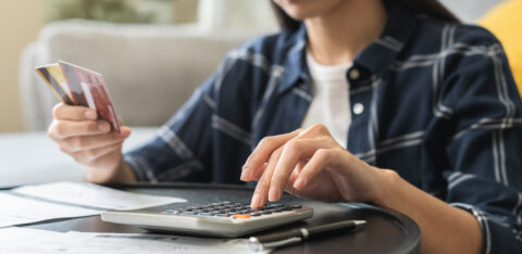 Close-up of a woman using a calculator while holding credit cards, with bills and paperwork spread out on a table.