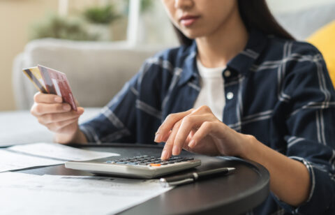 Close-up of a woman using a calculator while holding credit cards, with bills and paperwork spread out on a table.