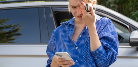 A woman standing next to a car looks stressed while holding her forehead, car keys, and a smartphone.