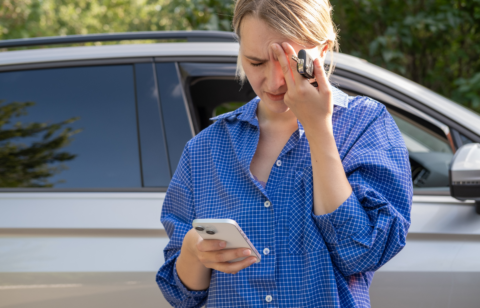 A woman standing next to a car looks stressed while holding her forehead, car keys, and a smartphone.