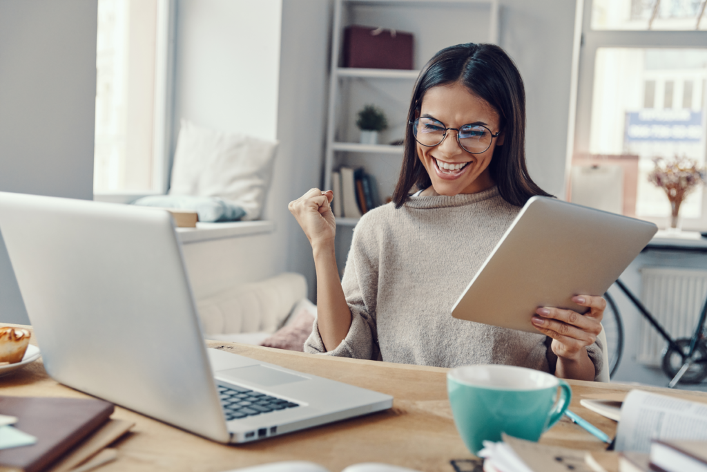 A woman smiling with excitement while holding a tablet and sitting at a desk with a laptop, coffee mug, and open books.