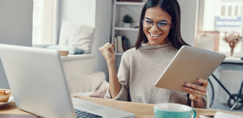 A woman smiling with excitement while holding a tablet and sitting at a desk with a laptop, coffee mug, and open books.