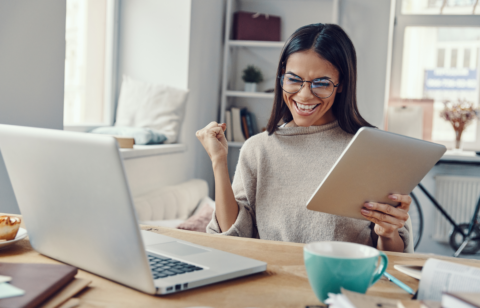 A woman smiling with excitement while holding a tablet and sitting at a desk with a laptop, coffee mug, and open books.