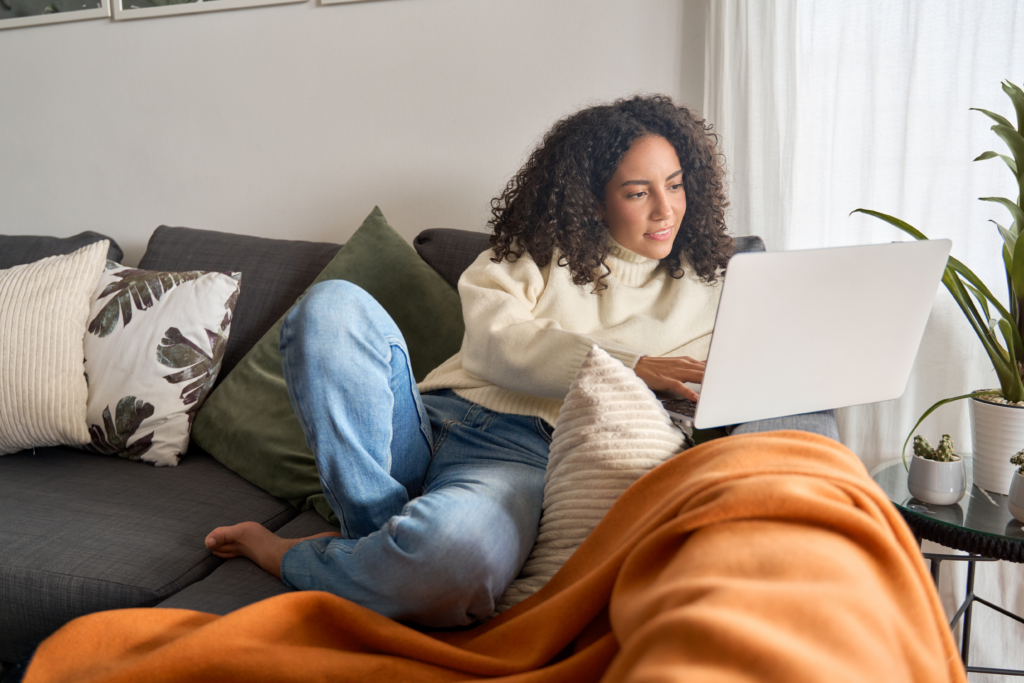 BYoung woman relaxing on a couch and working on a laptop, surrounded by pillows and cozy home decor.