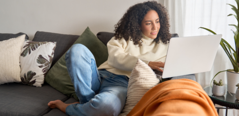 BYoung woman relaxing on a couch and working on a laptop, surrounded by pillows and cozy home decor.