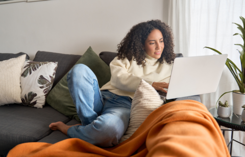 BYoung woman relaxing on a couch and working on a laptop, surrounded by pillows and cozy home decor.