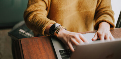 Person in a yellow sweater typing on a laptop at a wooden desk, with a bracelet on their wrist.