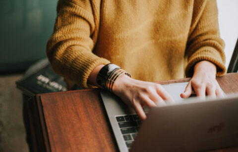 Person in a yellow sweater typing on a laptop at a wooden desk, with a bracelet on their wrist.