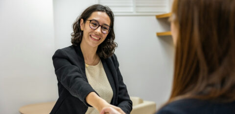 Smiling woman in professional attire shakes hands with another person across a desk in an office setting.