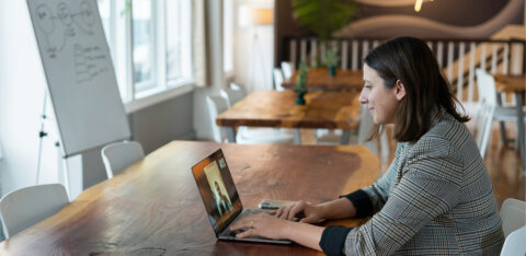 Professional woman in a plaid blazer sitting at a wooden table, smiling during a video call on her laptop in a modern, well-lit workspace.
