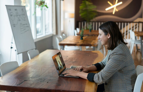 Professional woman in a plaid blazer sitting at a wooden table, smiling during a video call on her laptop in a modern, well-lit workspace.