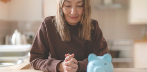 Middle-aged woman sitting at a kitchen table, looking thoughtfully at a blue piggy bank and calculator in front of her.