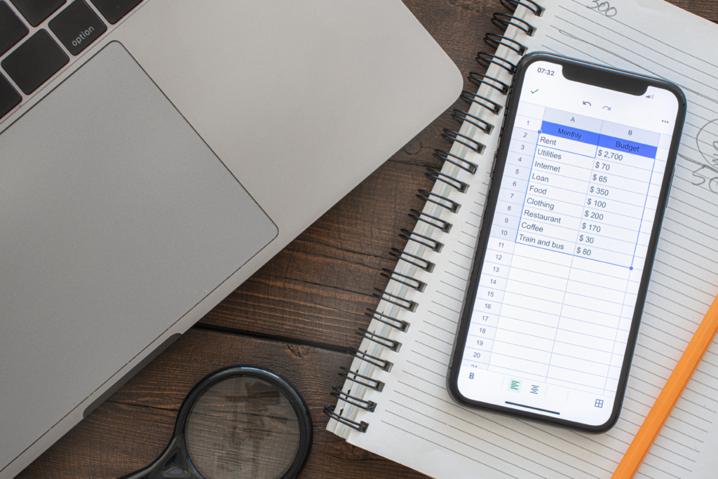 Smartphone displaying a monthly budget spreadsheet next to a laptop and notebook