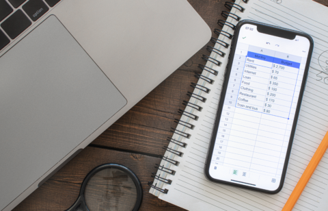 Smartphone displaying a monthly budget spreadsheet next to a laptop and notebook