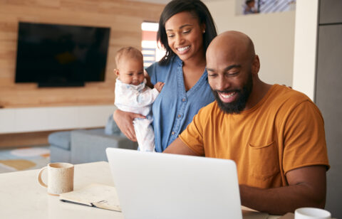 Smiling couple with a baby looking at a laptop together at home, appearing happy and engaged.