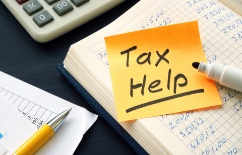 Close-up of a notebook with financial calculations, a yellow sticky note labeled "Tax Help," a white marker, calculator, and documents on a dark desk.