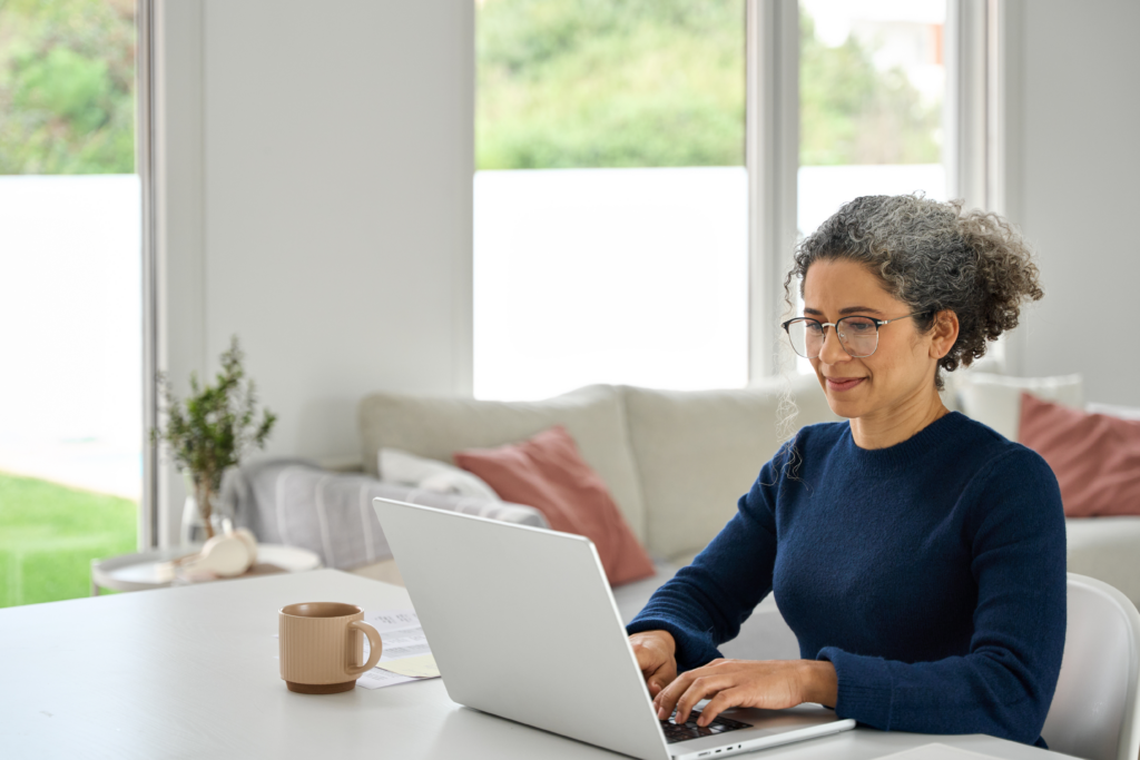 Middle-aged woman working on a laptop at home, smiling slightly while seated at a bright, modern table with a coffee mug nearby.