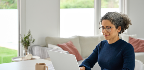 Middle-aged woman working on a laptop at home, smiling slightly while seated at a bright, modern table with a coffee mug nearby.