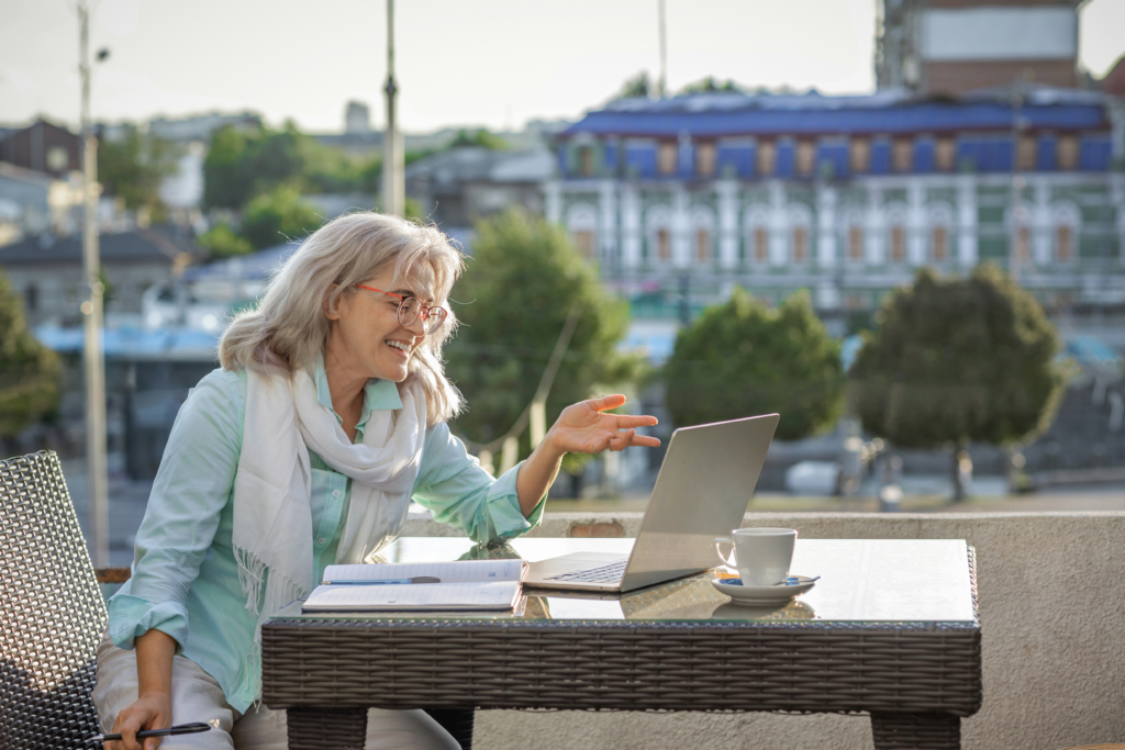 A smiling older woman sits at an outdoor café table, having a video call on her laptop with a notebook and coffee beside her.