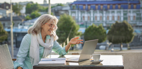 A smiling older woman sits at an outdoor café table, having a video call on her laptop with a notebook and coffee beside her.