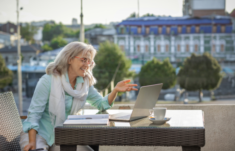 A smiling older woman sits at an outdoor café table, having a video call on her laptop with a notebook and coffee beside her.