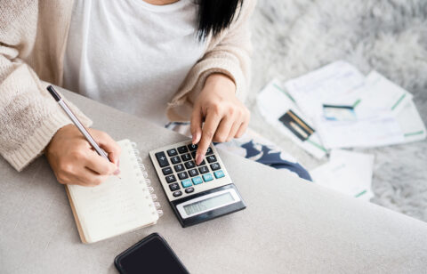 Woman calculating expenses with a large calculator and taking notes in a notebook, surrounded by bills and a smartphone.