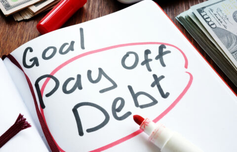 Notebook page with the words “Goal: Pay Off Debt” circled in red marker, surrounded by cash and a red pen.