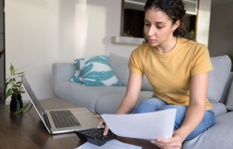 Young woman sitting on a couch, reviewing financial documents with a calculator and laptop on the coffee table.