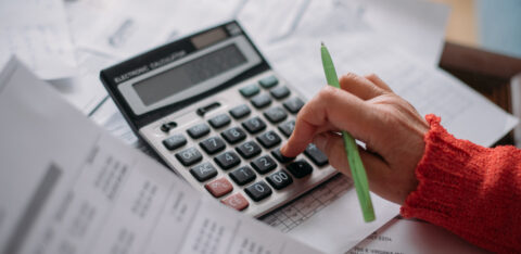 Person using a calculator while reviewing bills and financial documents, with a green pen in hand.