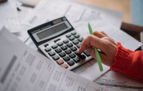 Person using a calculator while reviewing bills and financial documents, with a green pen in hand.