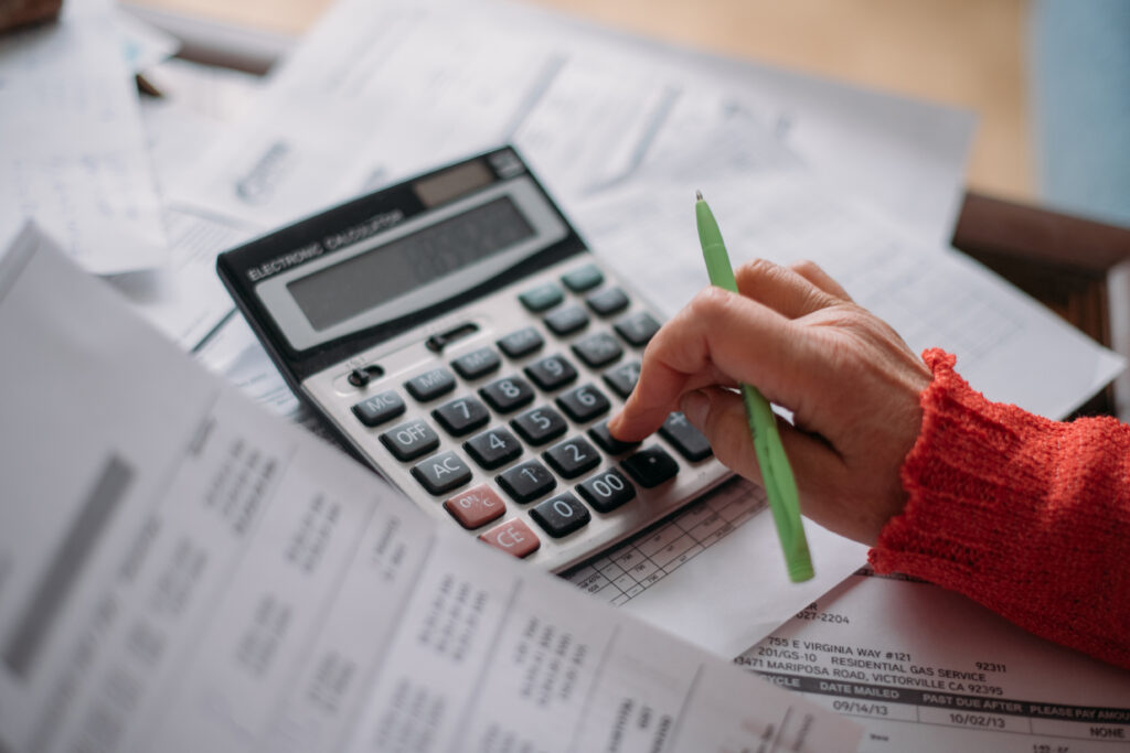 Close-up of a person using a calculator and reviewing bills, surrounded by financial documents and holding a green pen.